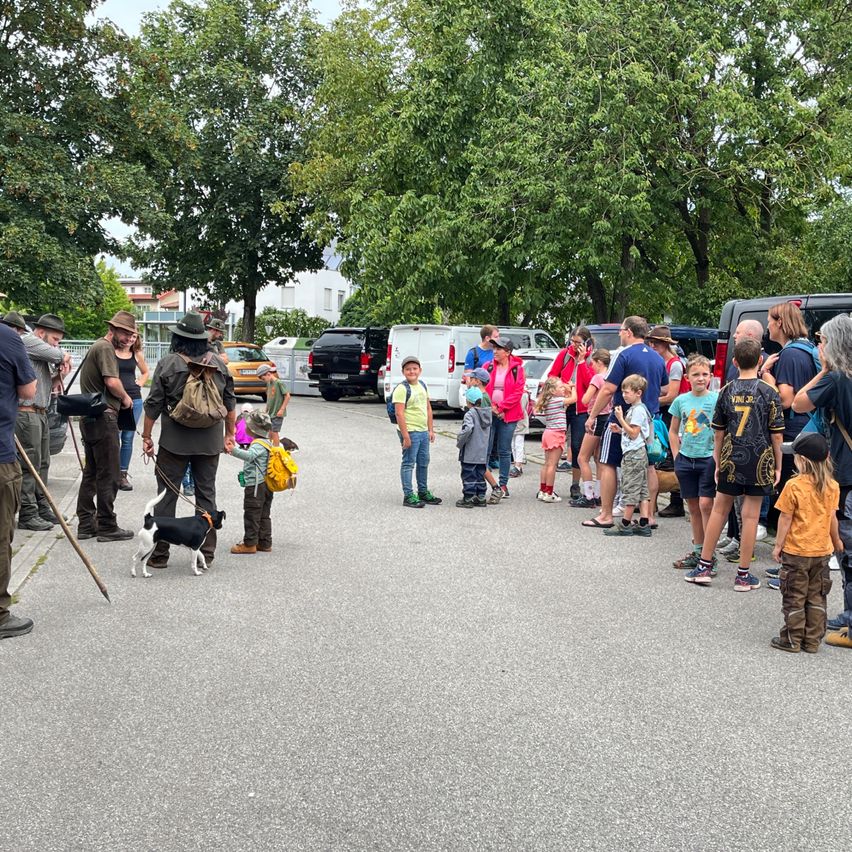 Menschen und Kinder stehen auf einem Parkplatz. Einige tragen Rucksäcke und Hüte. Ein Hund ist an der Leine. Ein weißer Van ist geparkt. Bäume sind im Hintergrund.