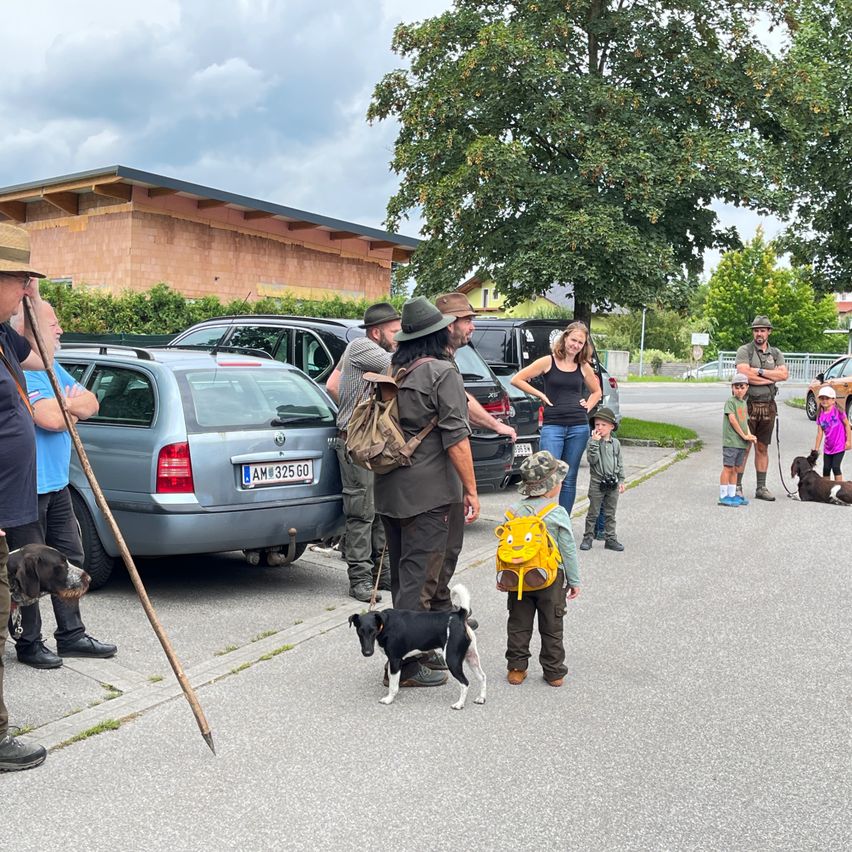 Eine Gruppe von Menschen und ihren Hunden versammelt sich neben geparkten Autos. Einige Personen tragen Hüte und haben Rucksäcke dabei. Ein Gebäude mit einem braunen Dach ist im Hintergrund.