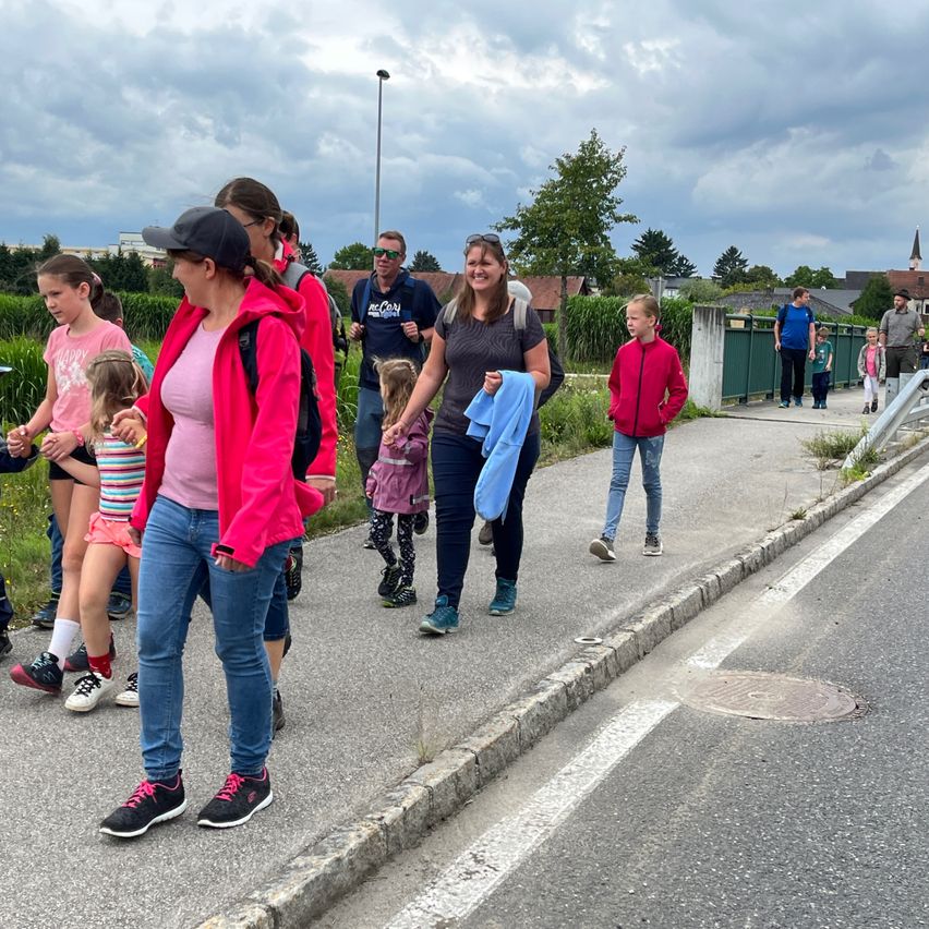 Eine Gruppe von Menschen, einschließlich Erwachsener und Kindern, geht auf dem Bürgersteig. Einige tragen Turnschuhe, während andere Rucksäcke tragen. Sie befinden sich in einem Außenbereich mit bewölktem Himmel.