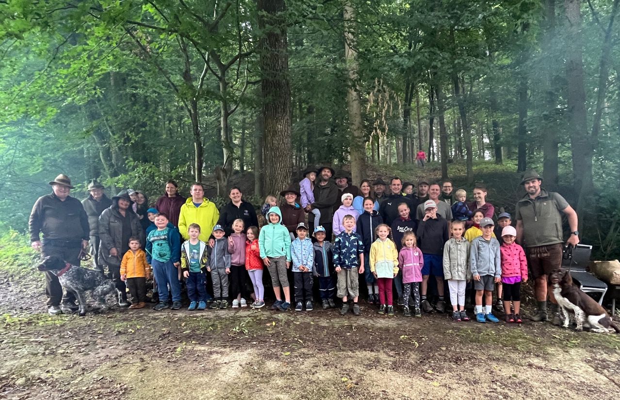 Eine Gruppe von Menschen, einschließlich Erwachsener und Kindern, steht in einem Wald. Einige tragen Hüte und Schuhe. Bäume umgeben das Gebiet.