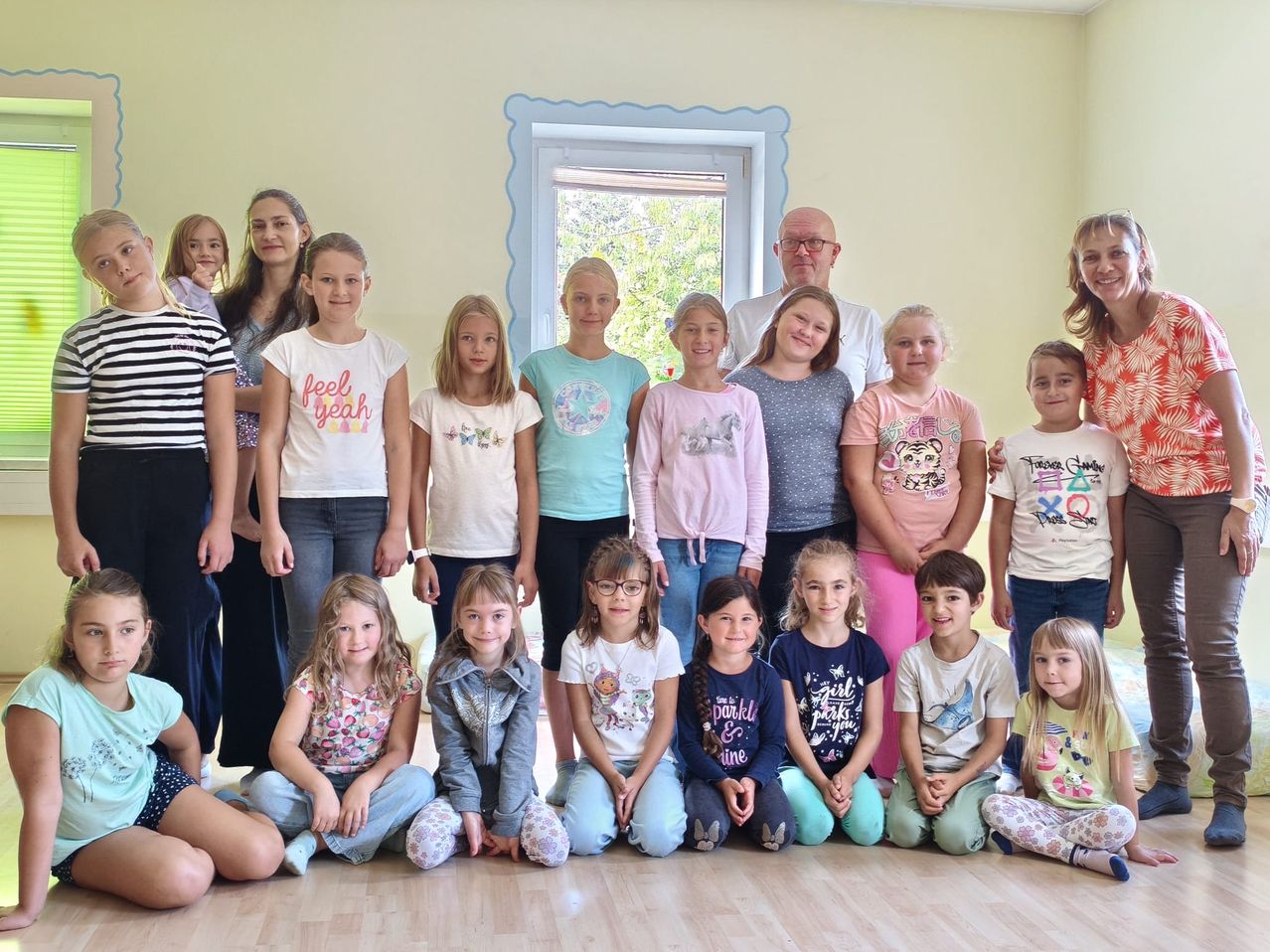 Eine Gruppe junger Mädchen und Jungen posiert für ein Foto in einem Raum mit gelber Wand und einem Fenster mit weißem Rahmen. Einige stehen, andere sitzen auf dem Boden. Sie alle lächeln.