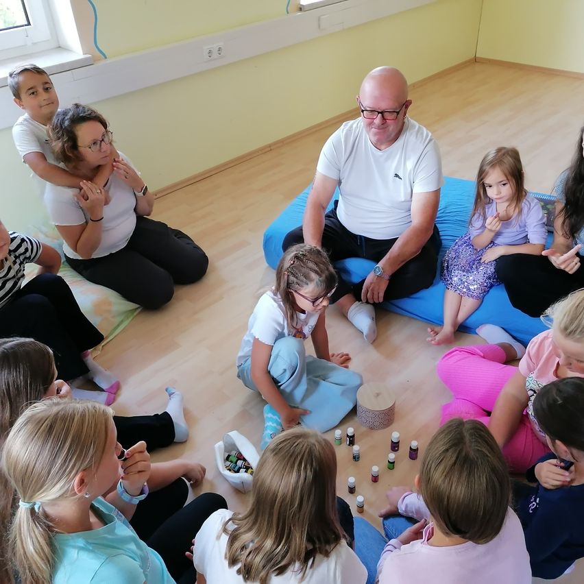 In einem Klassenzimmer sitzen eine Gruppe von Kindern und Erwachsenen im Kreis auf dem Boden. Ein Mann und eine Frau sitzen auf Kissen. Einige Kinder haben Flaschen und scheinen eine Aktivität durchzuführen.