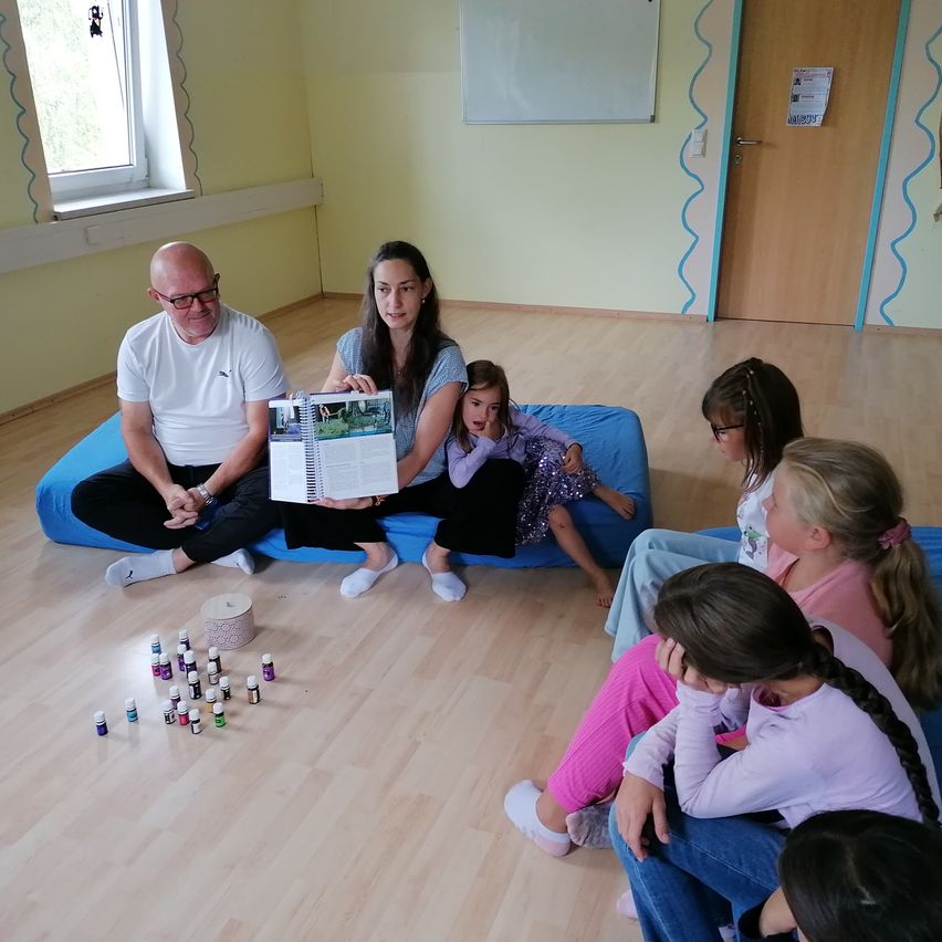 Eine Gruppe von Kindern sitzt auf blauen Kissen auf dem Boden. Eine erwachsene Frau liest ihnen ein Buch vor, während ein Mann neben ihr sitzt. Sie sind von kleinen Flaschen mit ätherischen Ölen umgeben. Der Raum hat Holzböden und eine gelbe Wand.