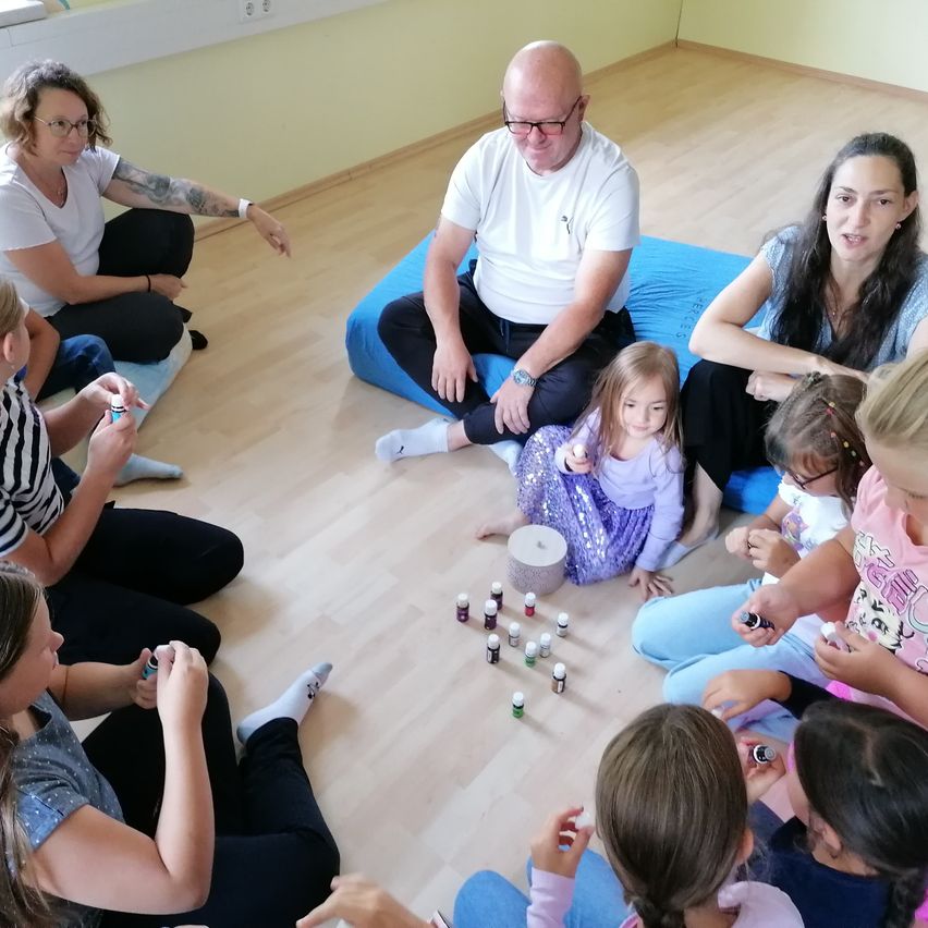 Eine Gruppe von Erwachsenen und Kindern sitzt in einem Kreis, wahrscheinlich in einem Klassenzimmer, mit ätherischen Ölen auf dem Boden. Ein Mann sitzt auf einem blauen Kissen, während ein Mädchen auf dem Boden sitzt. Einige der Kinder halten Flaschen.