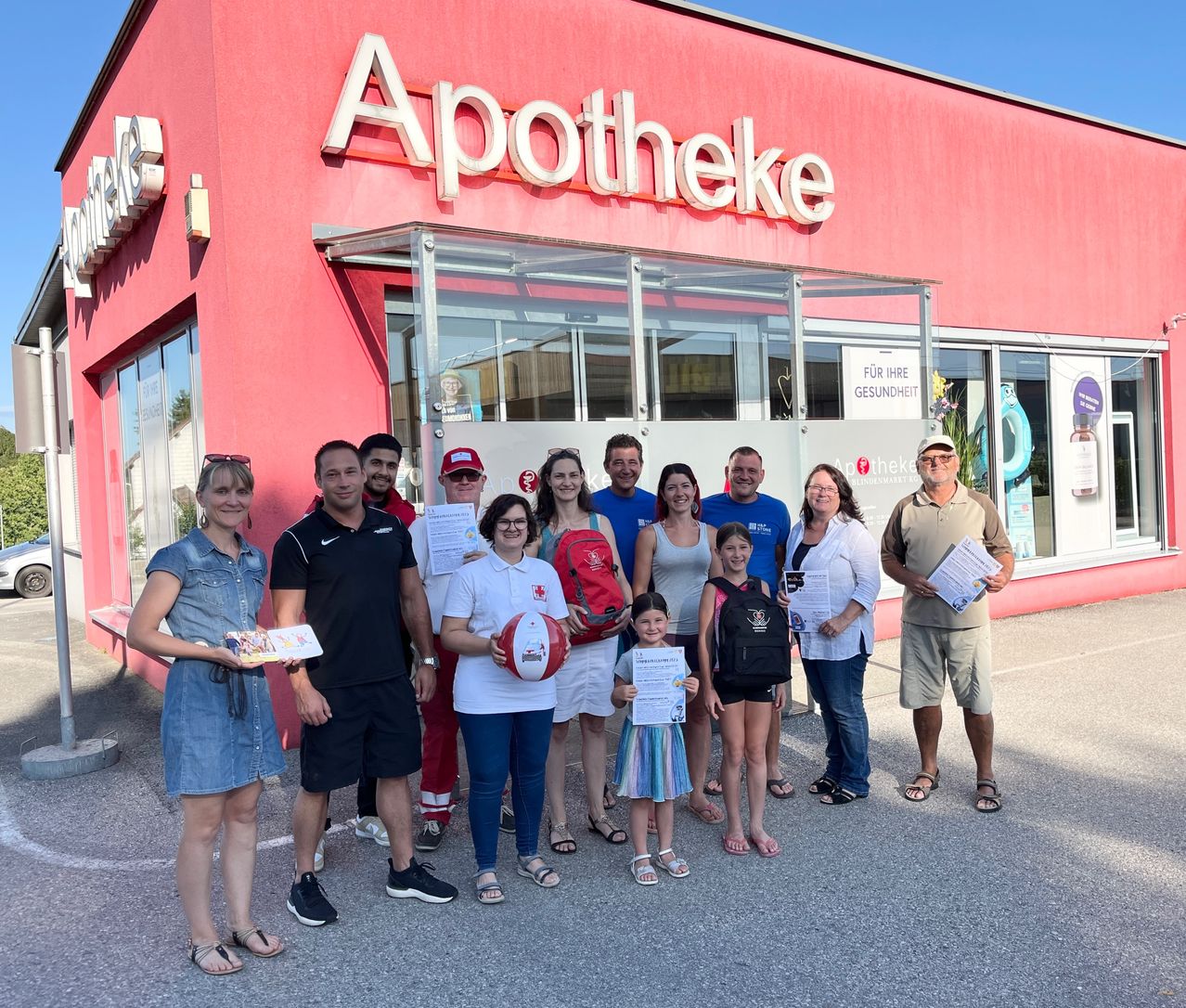 Eine Gruppe von Menschen steht vor einer Apotheke und lächelt für ein Foto. Papiere und Urkunden sind in ihren Händen. Die Apotheke ist leuchtend gefärbt, mit Glasfenstern und Beschilderung.