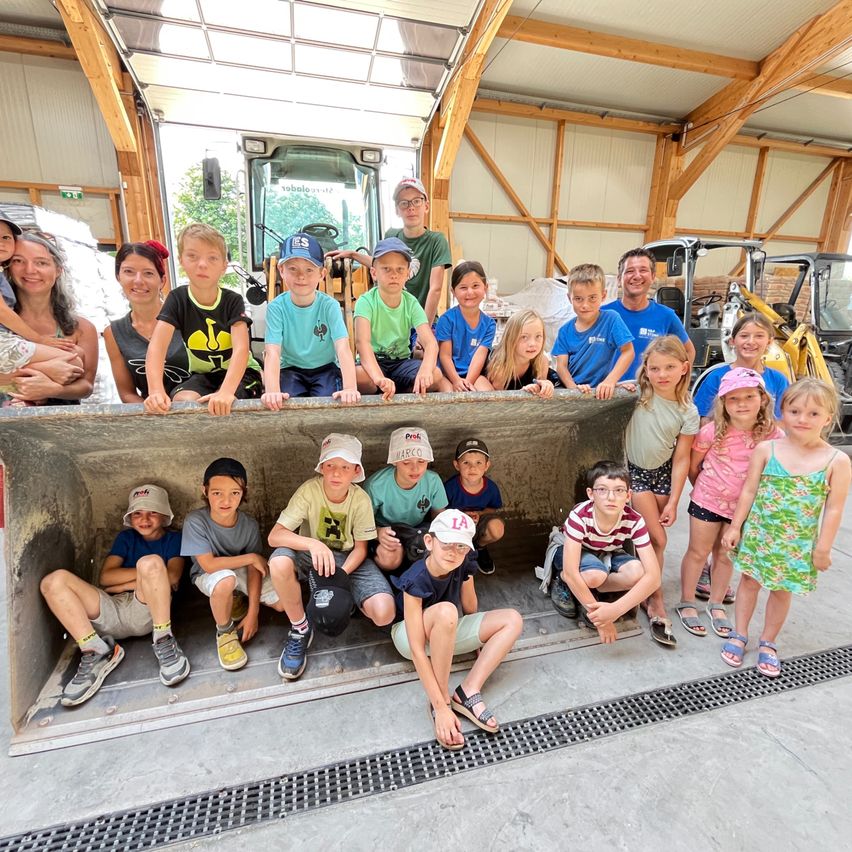 Eine Gruppe von Kindern und Erwachsenen, einige mit Mützen, posiert für ein Foto in einem Gebäude, wobei im Hintergrund Maschinen zu sehen sind.