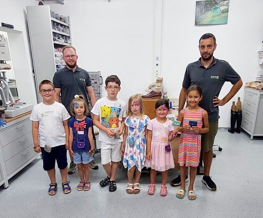 Eine Gruppe von Kindern und zwei Männern steht in einem Raum. Die Kinder haben Gesichtsbemalung. Sie halten eine Trophäe. Die Männer tragen Poloshirts. Hinter ihnen befinden sich Schränke und Regale.