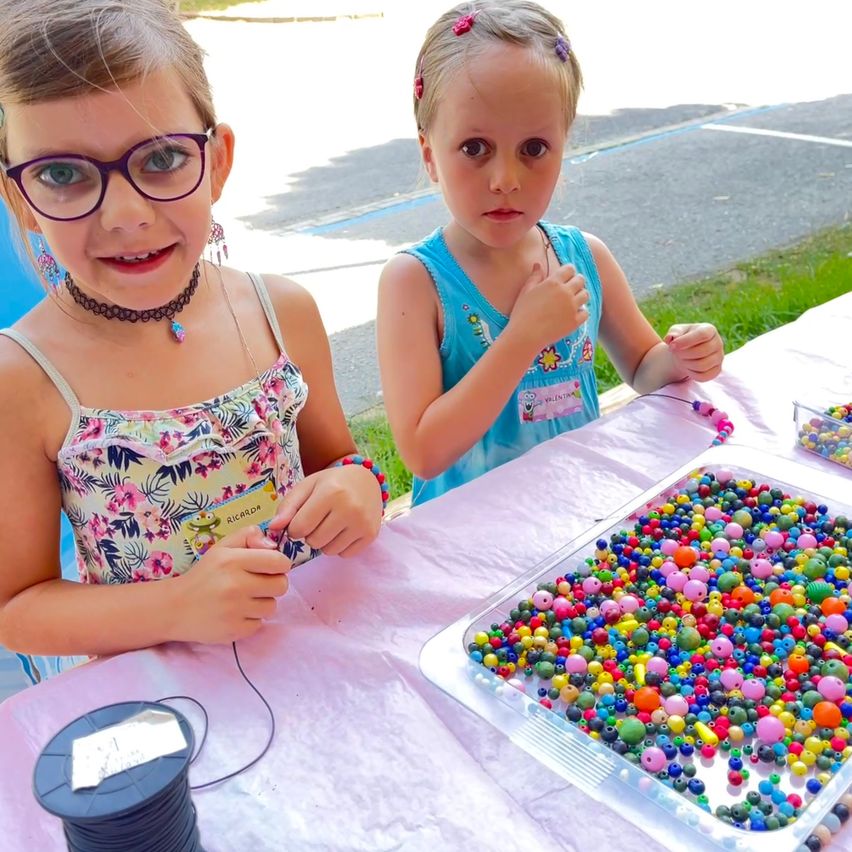 Zwei kleine Mädchen sitzen draußen an einem Tisch und beschäftigen sich mit einer Bastelaktivität. Ein Mädchen trägt eine Brille und eine Halskette und hält ein Stück Schnur in der Hand. Das andere Mädchen in einem blauen T-Shirt zeigt auf eine große Schale voller bunter Perlen. Ein Spule Faden liegt nebenan auf dem Tisch.