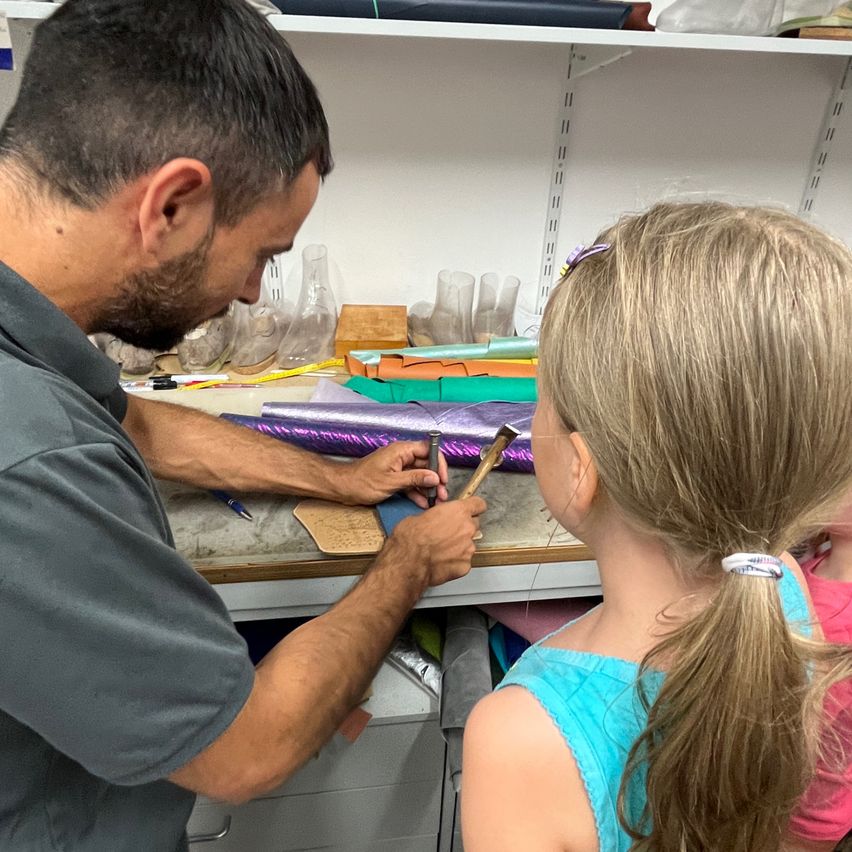 Ein Mann arbeitet an einem Bastelprojekt, während ein junges Mädchen ihn aufmerksam beobachtet. Der Mann benutzt einen Hammer und ein Stück lila Material.