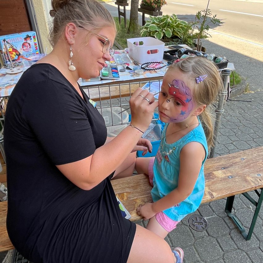 Eine Frau mit Brille malt ein Drachendesign auf das Gesicht eines jungen Mädchens. Sie sitzen auf einer Holzbank am Straßenrand.