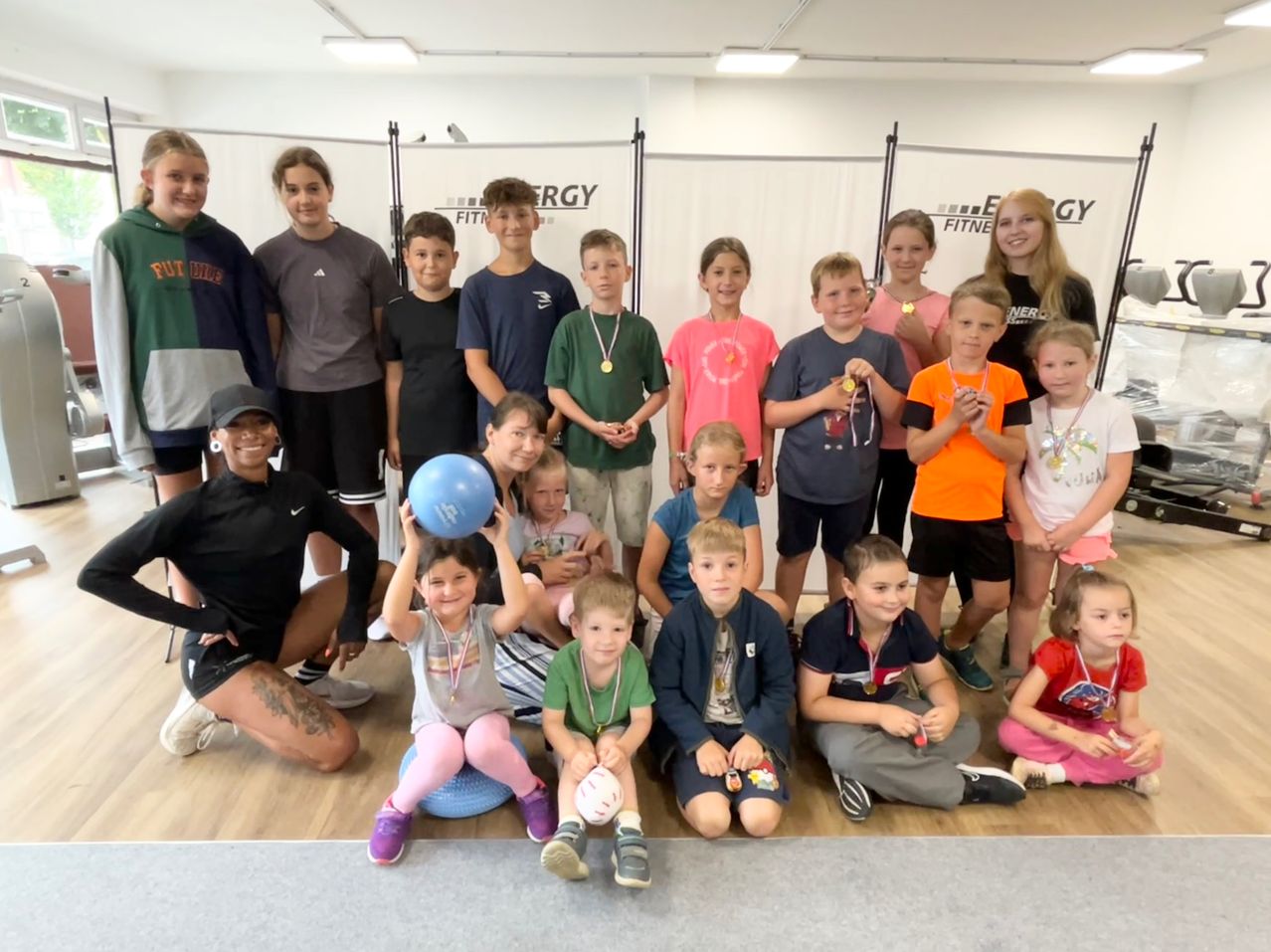 Eine Gruppe von Kindern und Erwachsenen posiert für ein Foto im Fitnessstudio, mit Medaillen. Ein Mädchen in rosa Hosen hält einen blauen Luftballon. Eine Frau mit einem Tattoo kniet neben einem Jungen mit einem Fußball.