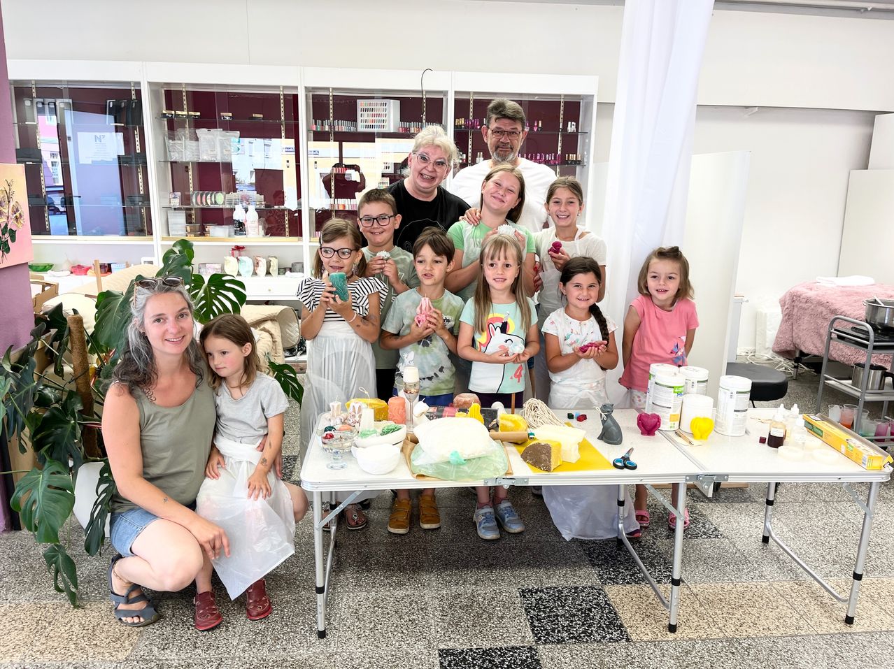 Eine Gruppe von Kindern und Erwachsenen versammelt sich um einen Tisch, lächelt und zeigt verschiedene Bastelmaterialien. Sie scheinen an einer Bastelaktivität beteiligt zu sein.