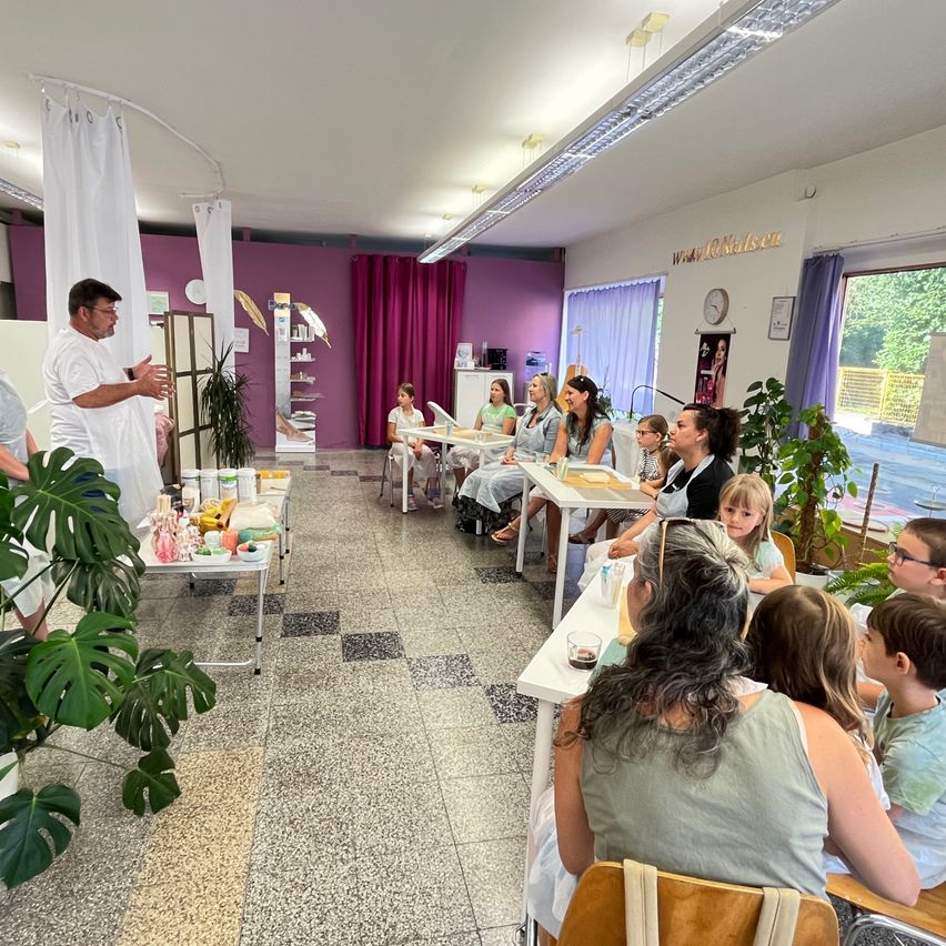 Eine Gruppe von Menschen, einschließlich Kindern, sitzt an Tischen in einem Raum mit lila Wänden. Ein Mann steht vorne, und verschiedene Gegenstände sind auf den Tischen. Es gibt Pflanzen und Vorhänge im Raum.