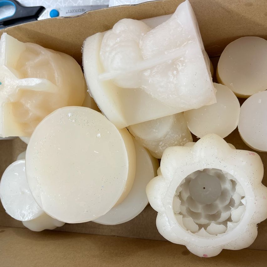 Ein Karton mit verschiedenen weißen Kerzenformen, darunter runde, blumenähnliche und zylindrische Formen. Die Kerzen haben eine glatte Textur und sind eng nebeneinander angeordnet.