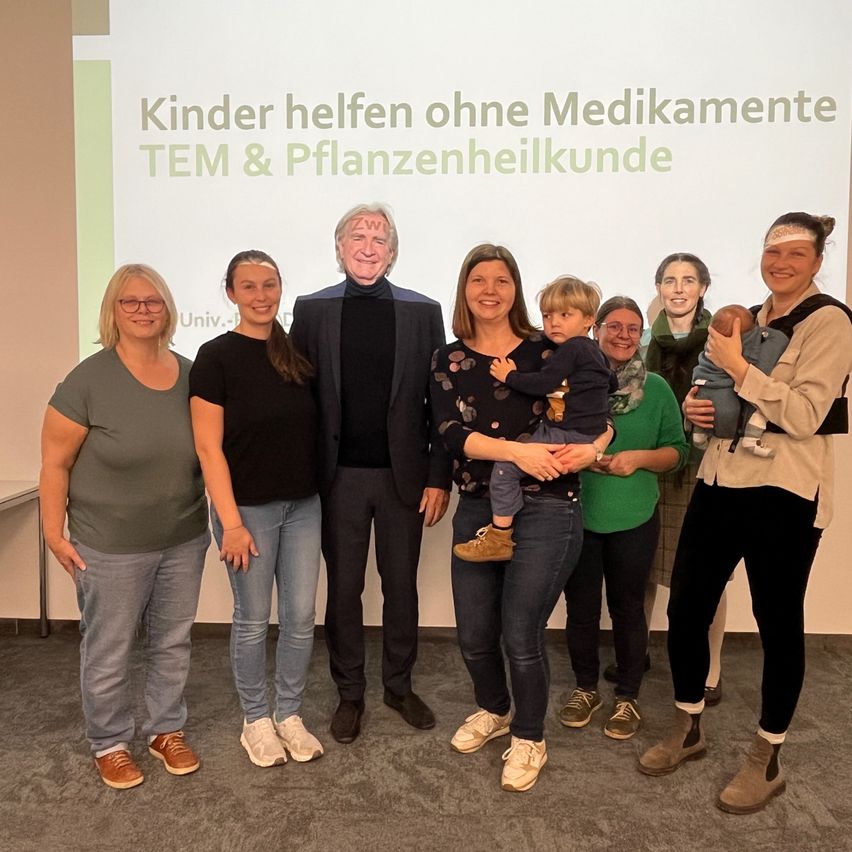 Eine Gruppe von Erwachsenen und Kindern steht lächelnd vor einer Präsentationsfolie. Die Folie besagt: 'Kinder helfen ohne Medikamente TEM & Pflanzenheilkunde'.