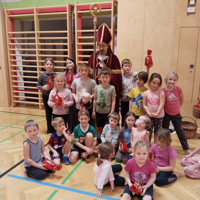 Eine Gruppe von Kindern posiert mit einem als Bischof gekleideten Mann. Sie halten rote Geschenktüten, und die Umgebung scheint ein Indoor-Spielbereich zu sein.