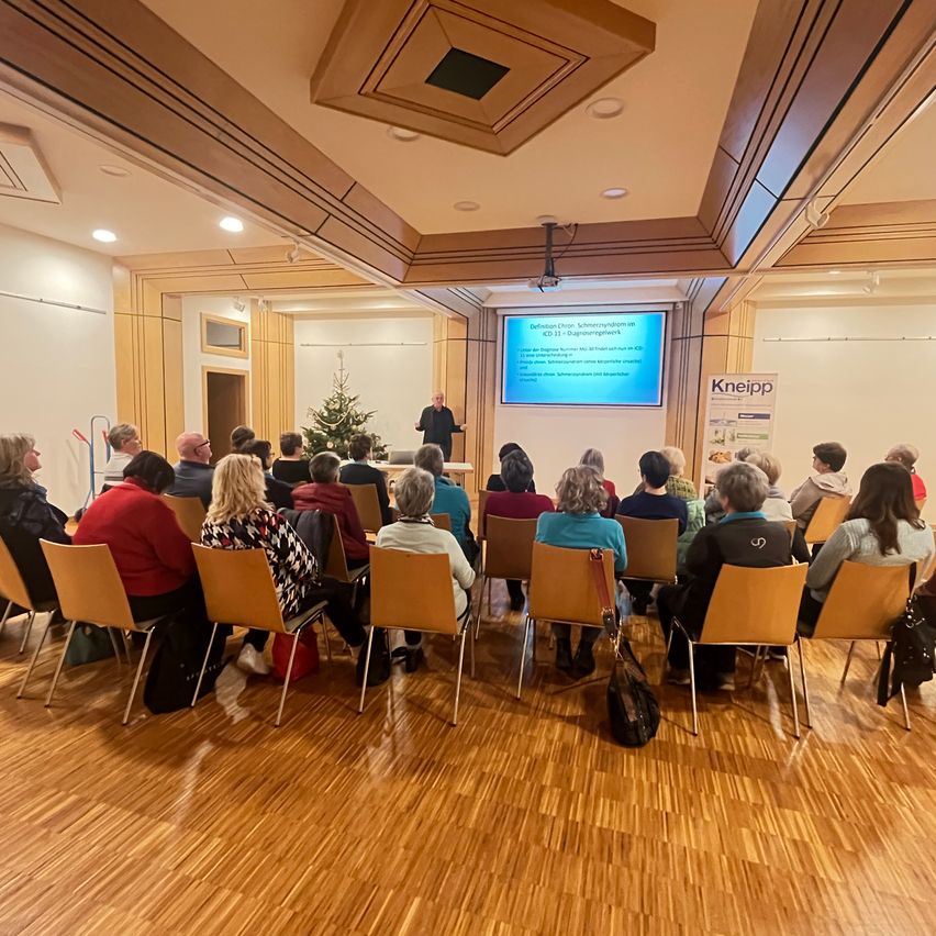 Eine Gruppe von Menschen sitzt in Stühlen vor einer Bühne, auf der ein Mann spricht, mit einer Projektionsleinwand im Hintergrund. Ein Weihnachtsbaum steht auf der Bühne.