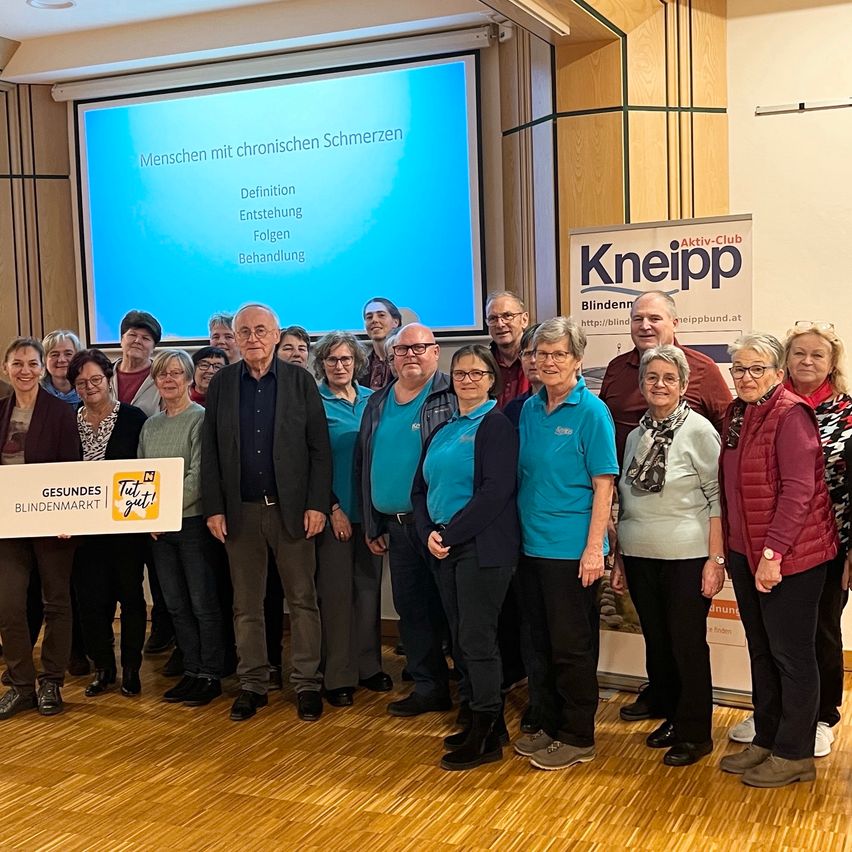 Eine Gruppe von Menschen in blauen Shirts, die vor einer Präsentationsleinwand stehen, mit einem Banner hinter ihnen, das 'Kneiipp' liest.