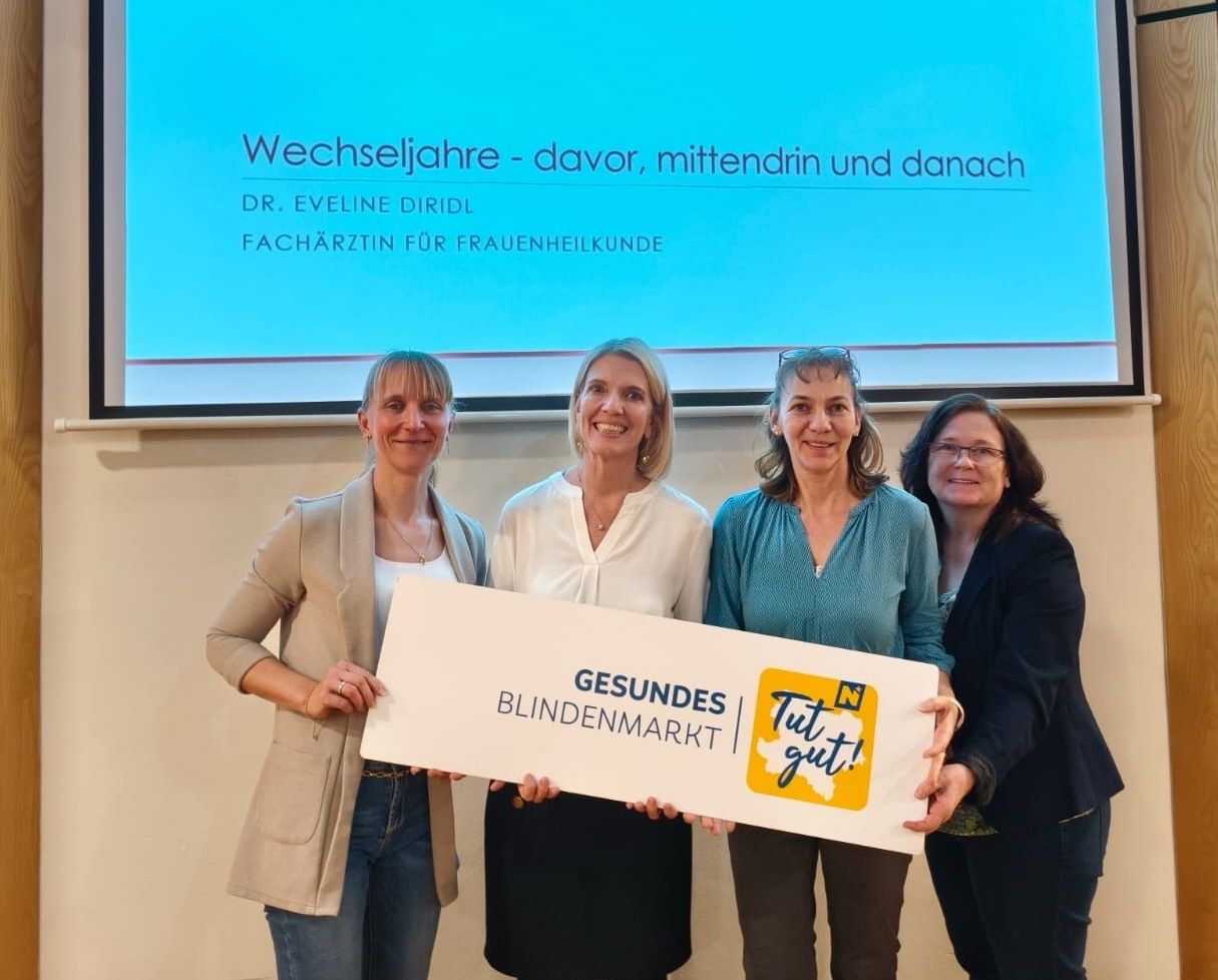 Vier Frauen stehen vor einer Leinwand. Sie halten ein großes Schild, auf dem 'Gesundes Blindenmarkt' steht. Dahinter ist eine Präsentationsfolie zu sehen, mit dem Titel 'Wechseljahre - davor, mittendrin und danach'.