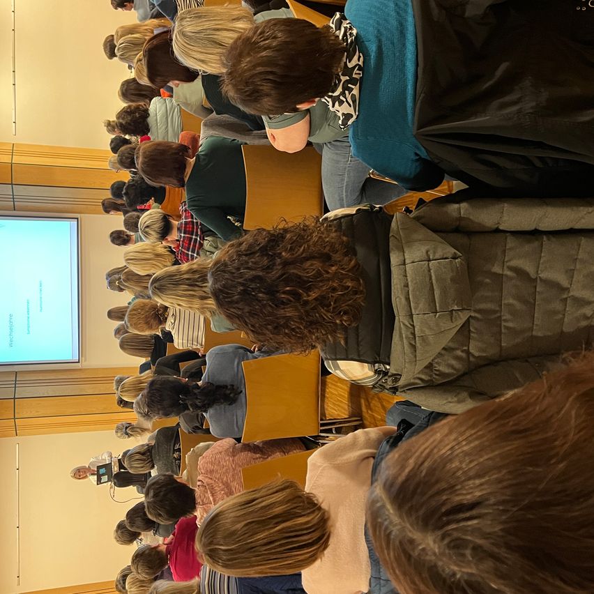 Ein Publikum sitzt in einem Hörsaal und schaut sich eine Präsentation auf einer Leinwand an. Eine Person steht an einem Podium am vorderen Ende des Raumes.