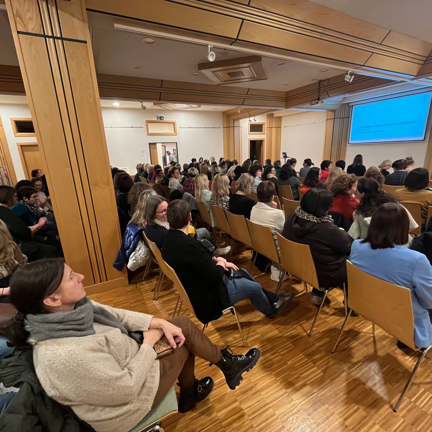 Ein überfüllter Raum mit vielen Stühlen und sitzenden Menschen, eine Projektionsleinwand an der Wand und eine offene Tür in der Ferne.