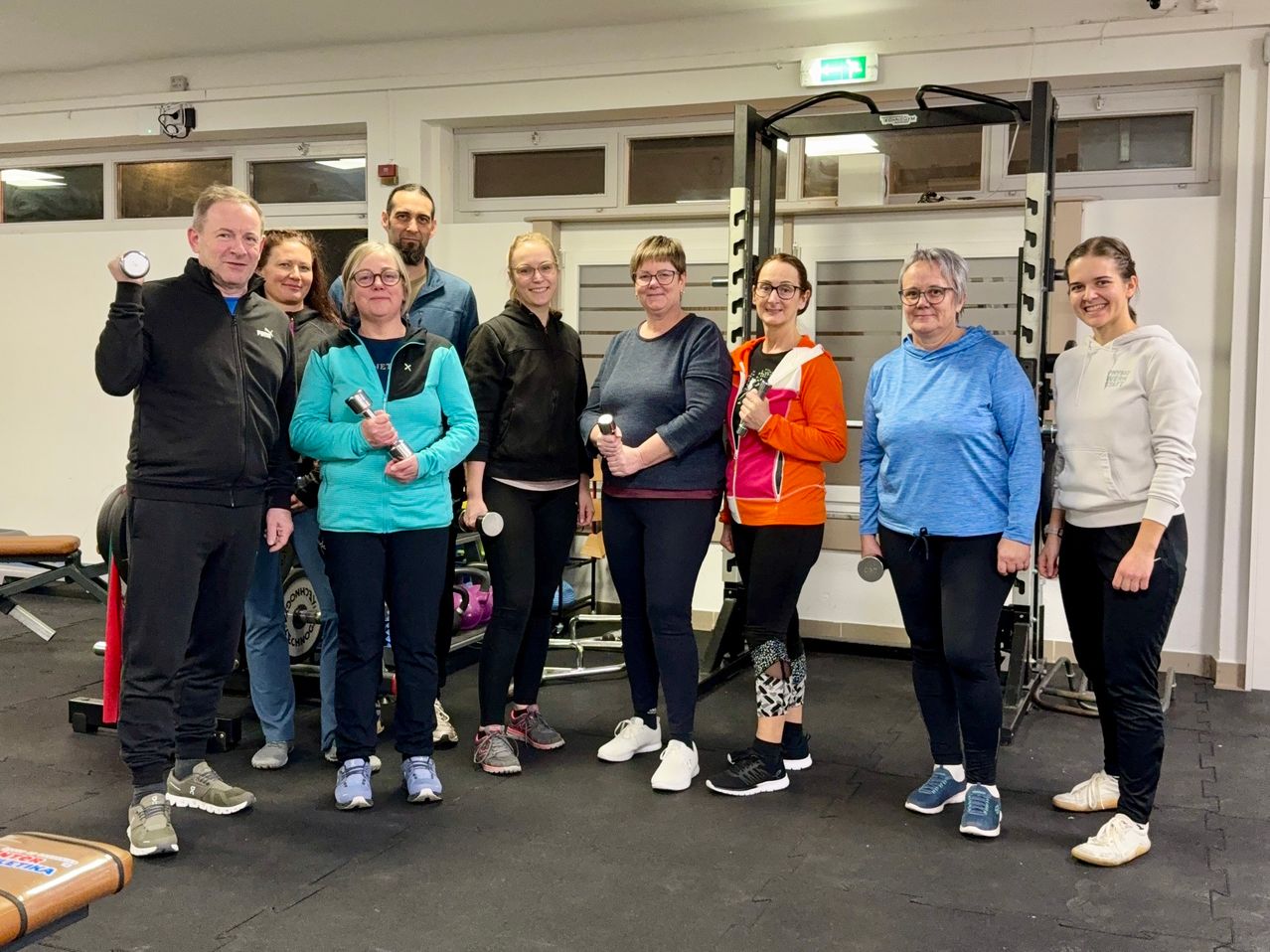 Eine Gruppe von Erwachsenen steht in einem Fitnessstudio und hält Hanteln. Sie lächeln und scheinen für ein Foto zu posieren. Im Hintergrund befinden sich verschiedene Trainingsgeräte.