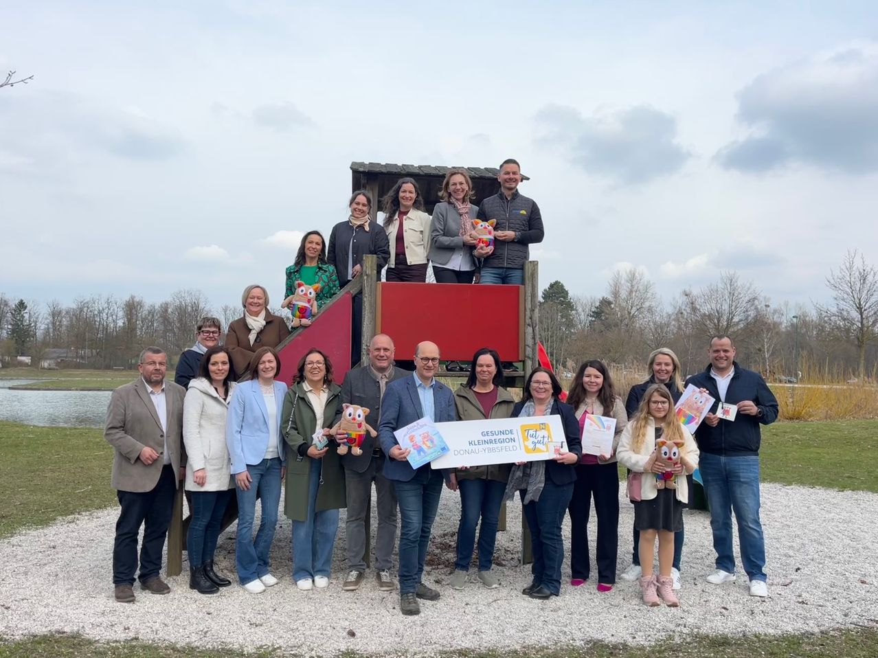 A group of people stands in front of a playground, holding certificates and stuffed toys. Behind them is a red structure with a sign. They seem to be celebrating an event.