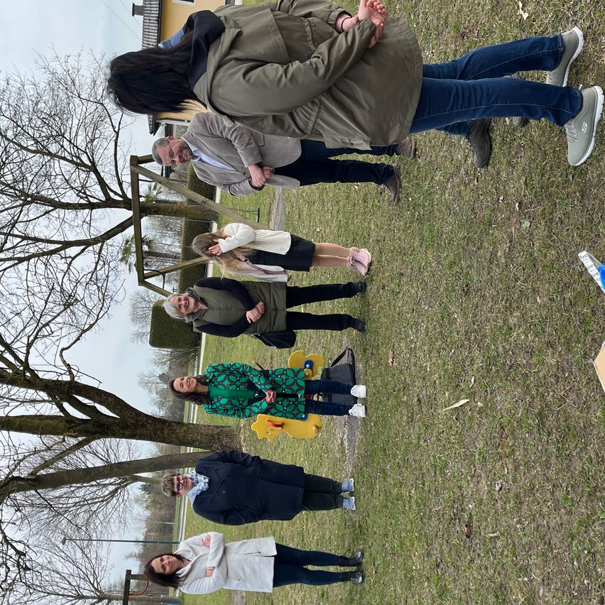 Seven people pose for a photo in an outdoor area, possibly a park, on a cloudy day. They are standing in a circle with a tree in the center.