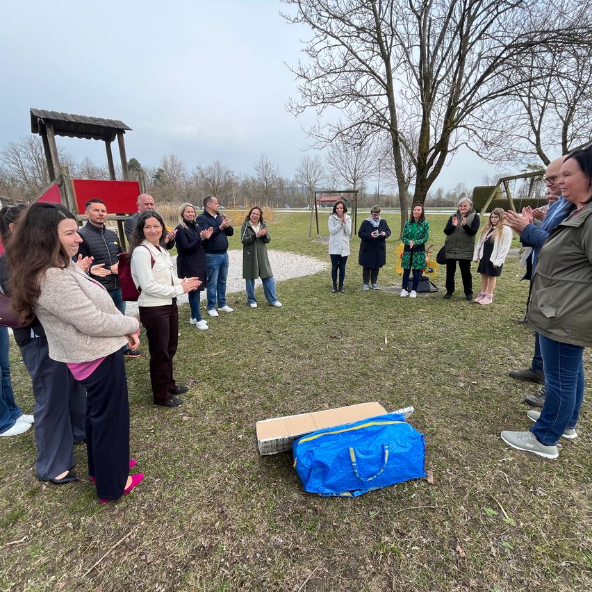 A group of people are gathered in a park, clapping and smiling. A blue bag and a cardboard box lie on the ground. Trees and a playground are visible in the background.