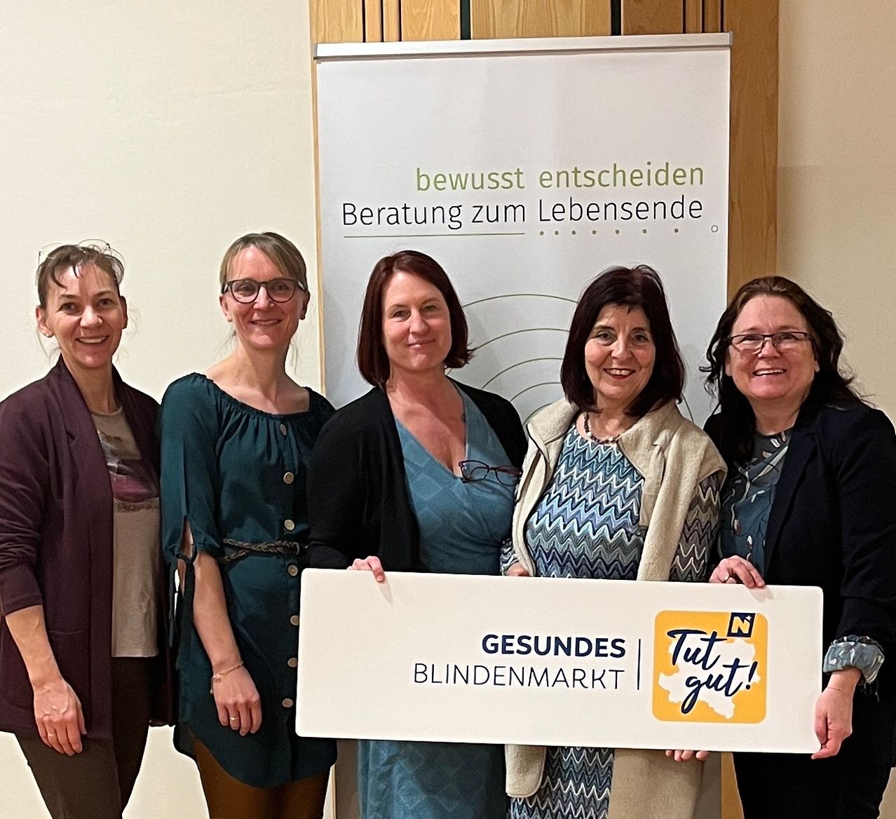 Fünf Frauen stehen vor einem weißen Banner. Das Banner heißt 'Gesundes Blindenmarkt'. Die Frauen halten ein Schild mit der Aufschrift 'Gut'. Sie alle lächeln. Hinter ihnen ist eine Holzwand.