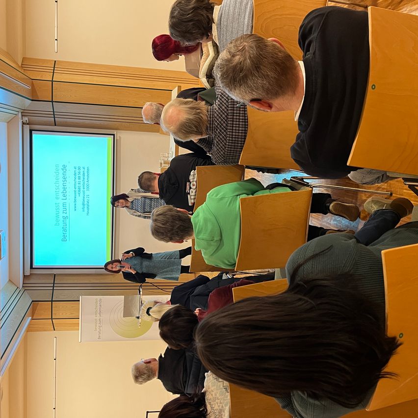 Eine Indoor-Konferenz mit einer Projektionsleinwand im Hintergrund. Zwei Frauen, eine mit einem Mikrofon, stehen vor der Leinwand. Eine Menschenmenge sitzt in Stühlen und hört zu.