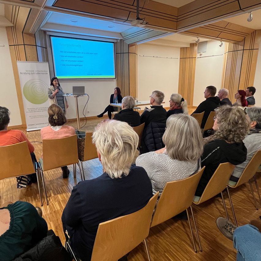 Eine Frau präsentiert an einem Rednerpult in einem Raum mit einem großen Bildschirmprojektor. Mehrere Menschen sitzen vor ihr auf Stühlen. Der Raum ist gut beleuchtet.