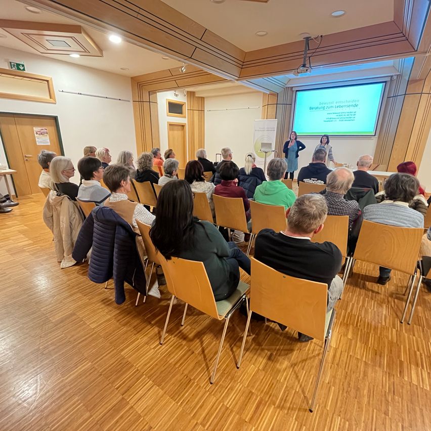 Eine Gruppe von Menschen sitzt in Stühlen in einem Raum und beobachtet eine Präsentation auf einer Projektionsleinwand. Zwei Frauen stehen vor der Leinwand und halten wahrscheinlich eine Präsentation. Der Raum hat Holzböden und weiße Wände.