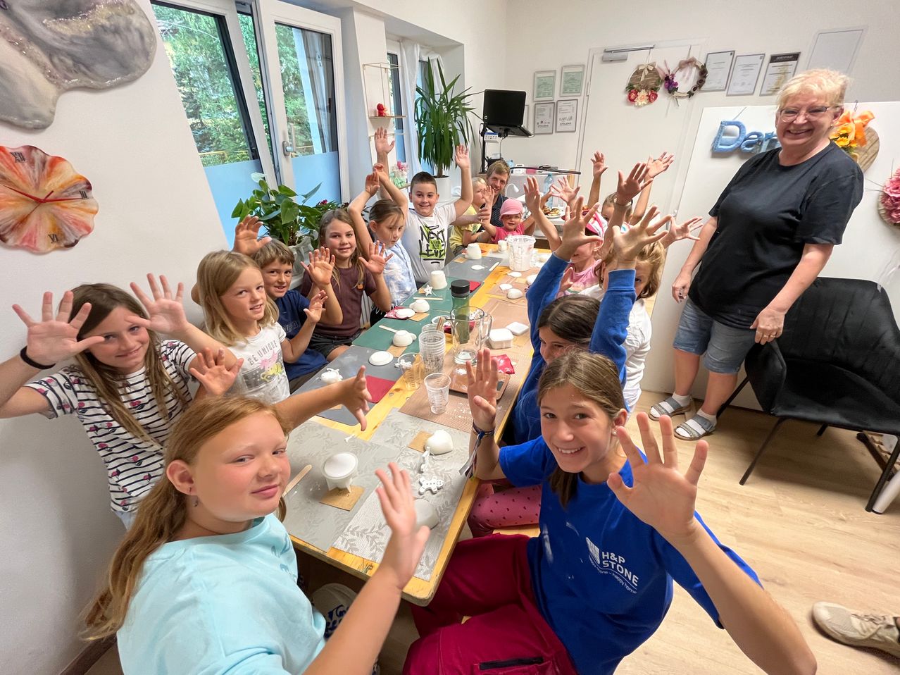 Eine Gruppe von Kindern und einer Frau sind in einem Raum, lächeln und heben ihre Hände. Sie sitzen um einen Tisch mit Tassen, Tellern und anderen Gegenständen. Die Frau steht hinter dem Tisch. Der Raum hat eine weiße Wand, ein Fenster und eine Pflanze.