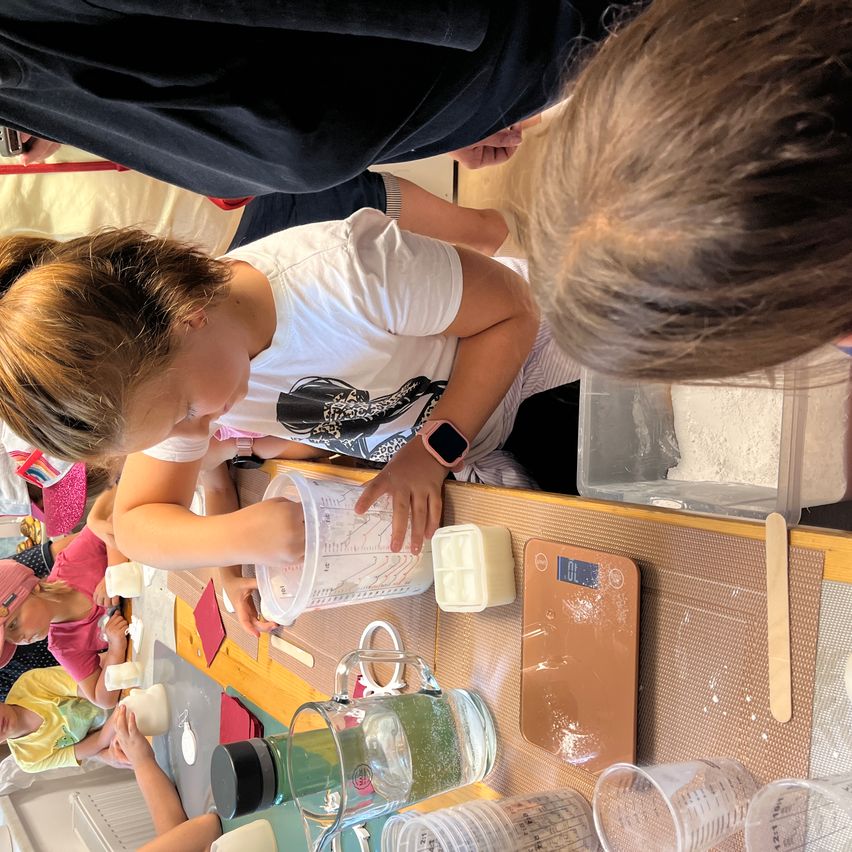 Eine Gruppe von Kindern ist in eine Backaktivität verwickelt, wobei ein Mädchen Zutaten in einen Messbecher gießt. Andere Kinder sind in der Nähe, und verschiedene Backwerkzeuge und Zutaten sind auf dem Tisch.