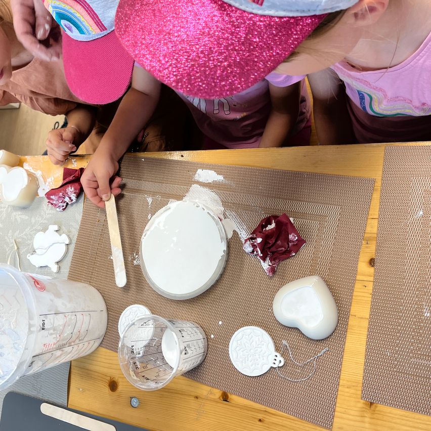 Eine Gruppe von Kindern arbeitet an einem Bastelprojekt an einem Tisch. Sie haben verschiedene Materialien verteilt, einschließlich eines Messbechers, einer Rührschüssel und eines herzförmigen Objekts. Ein Kind hält einen Stock und scheint etwas zu mischen.