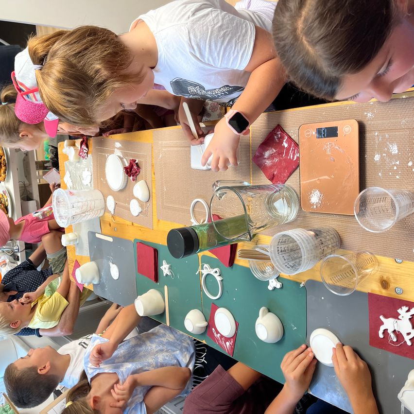 Eine Gruppe von Kindern ist um einen Tisch versammelt und beschäftigt sich mit einer Bastelaktivität. Sie verwenden verschiedene Werkzeuge und Materialien, darunter Becher, Teller und eine digitale Waage. Die Kinder tragen Freizeitkleidung und scheinen die Aktivität zu genießen.