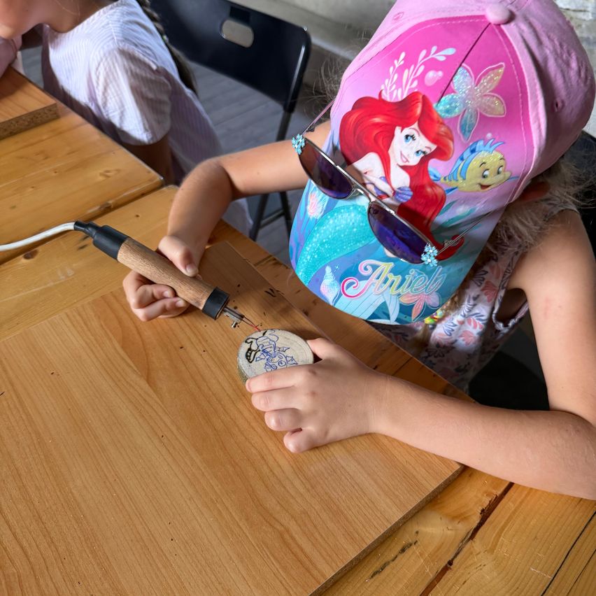 Ein kleines Mädchen mit einer rosa Ariel-Kappe und Sonnenbrille benutzt ein Holzbrandwerkzeug auf einem kleinen Holzkreis.