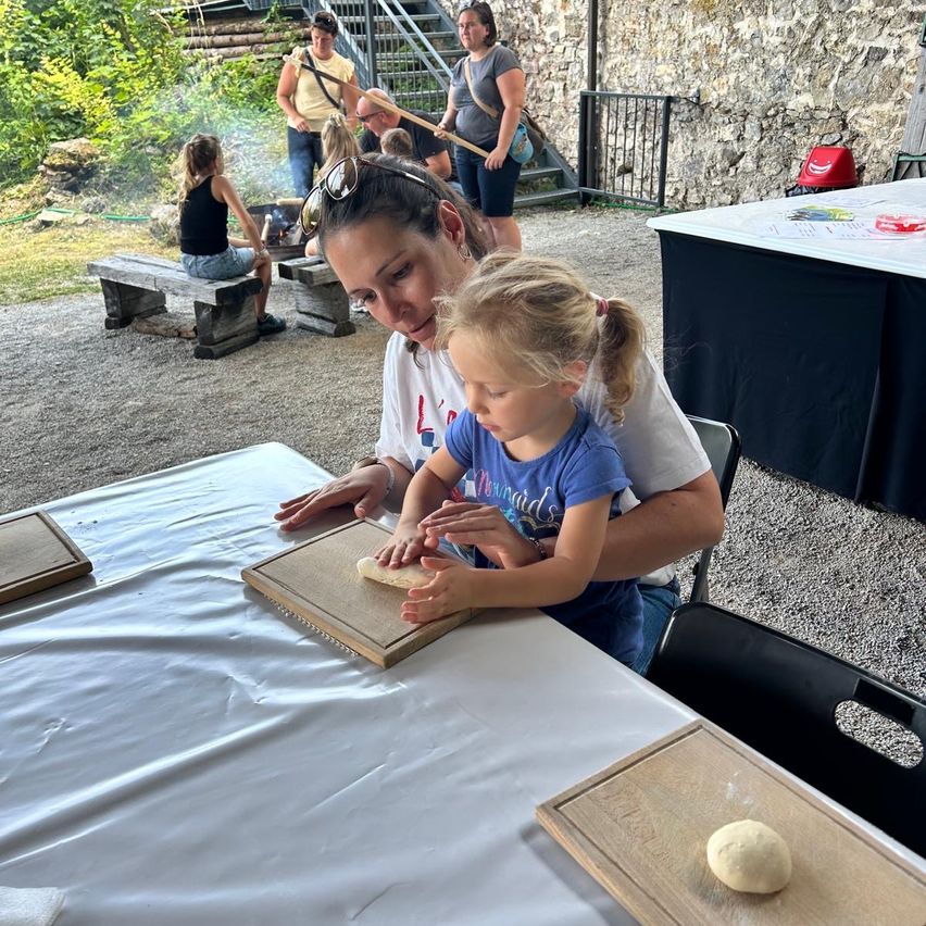 Eine Frau und ein junges Mädchen sitzen an einem Tisch und kneten Teig auf einem Holzbrett. Im Hintergrund sitzen eine Gruppe von Menschen auf einer Bank und stehen in der Nähe einer Steinmauer.