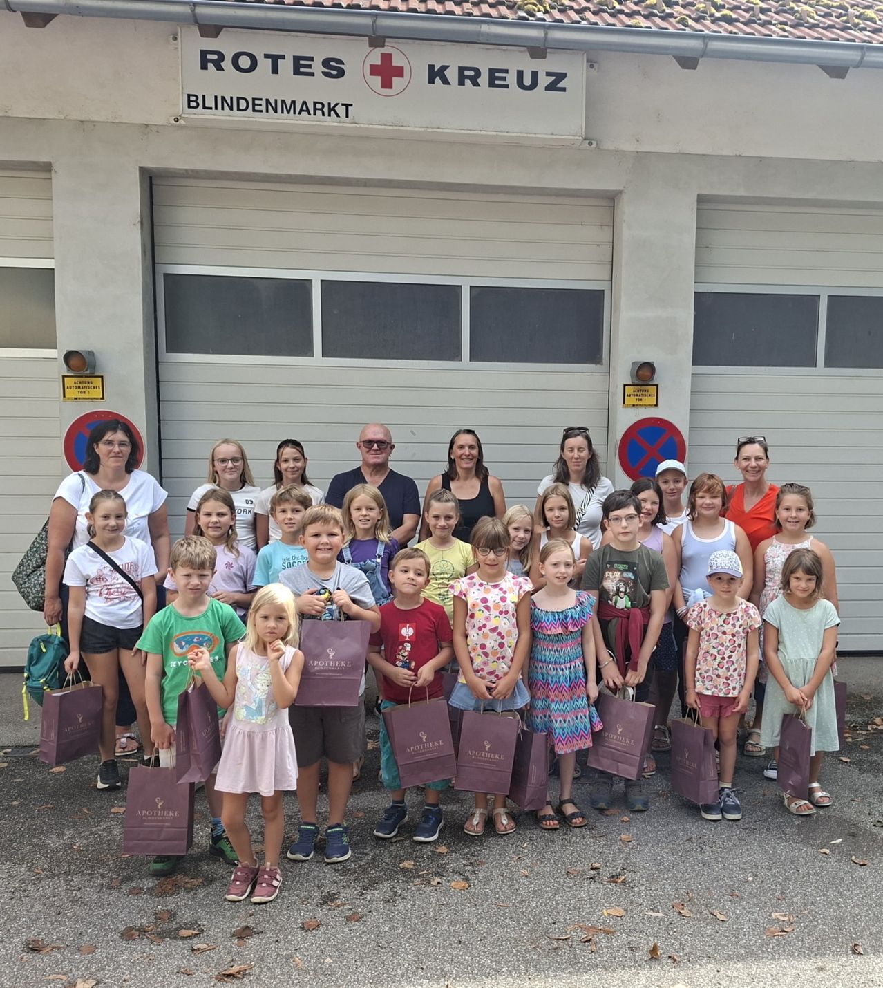 Eine Gruppe von Kindern und Erwachsenen posiert für ein Foto vor einem Gebäude mit einem Schild, das Rotes Kreuz Blindenmarkt besagt. Sie halten alle lila Taschen.