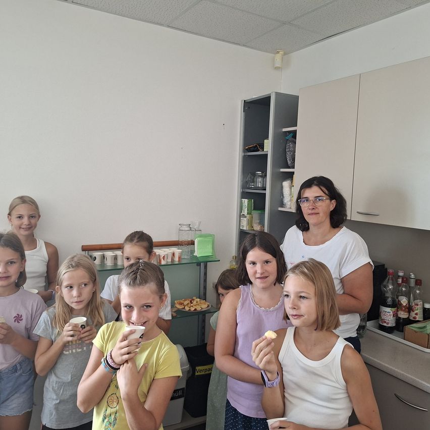 Eine Gruppe junger Mädchen und eine Frau sind in einer Küche, lächeln und essen Snacks. Die Frau steht hinter ihnen, und es gibt Regale und einen Tisch mit Essen im Hintergrund.
