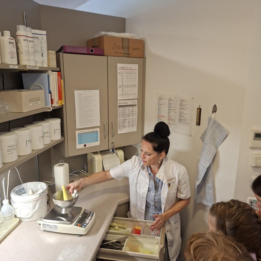 Eine Frau in einem Laborkittel demonstriert eine Prozedur für eine Gruppe von Kindern in einem Labor. Sie benutzt eine Waage und einen Behälter mit gelber Flüssigkeit.