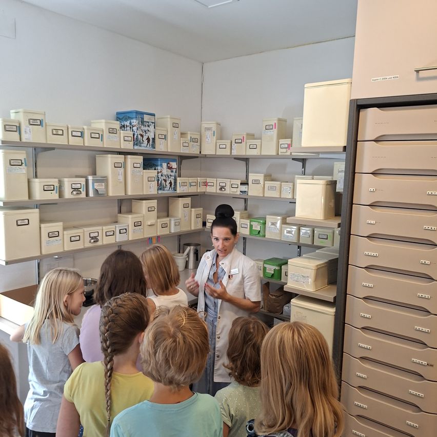 Eine Frau in einem weißen Laborkittel erklärt etwas einer Gruppe von Kindern in einem Raum, der mit ordentlich organisierten Aufbewahrungsboxen an Regalen gefüllt ist.