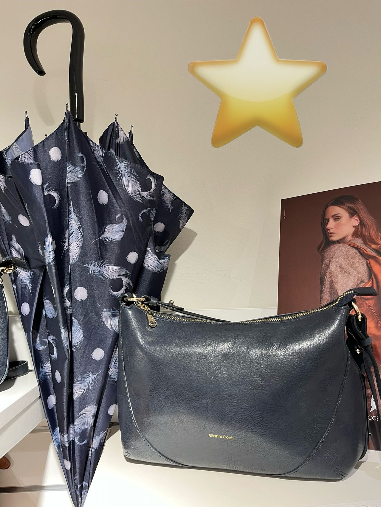 Eine schwarze Lederhandtasche mit goldenen Reißverschlüssen hängt neben einem blauen, federgemusterten Regenschirm. Ein leuchtender gelber Stern hängt darüber, und ein Bild einer Frau ist rechts.