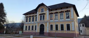 Ein gelbes und weißes Gebäude mit vielen Fenstern und einem schwarzen Dach steht vor einer Straße, mit einem Rasenbereich und einer Straßenlaterne davor.