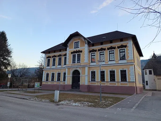 Ein gelbes und weißes Gebäude mit vielen Fenstern und einem schwarzen Dach steht vor einer Straße, mit einem Rasenbereich und einer Straßenlaterne davor.