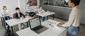 Eine Lehrerin steht vor einem Klassenzimmer mit drei Schülern an Schreibtischen. Ein Laptop ist auf dem Schreibtisch mit einem Schulzeitplan geöffnet. Die Schüler haben Bücher und Notizbücher vor sich. Das Klassenzimmer hat eine Ankündigungstafel und eine Glaswand.