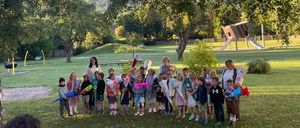 Eine Gruppe von Kindern steht auf einem Grasfeld und hält Blumensträuße in den Händen. Eine Frau steht unter ihnen, und sie alle scheinen für ein Foto zu posieren. Dahinter befindet sich ein Spielplatz mit Rutschen.