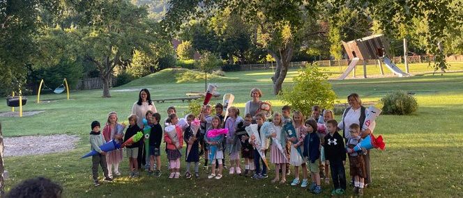 Eine Gruppe von Kindern steht auf einem Grasfeld und hält Blumensträuße in den Händen. Eine Frau steht unter ihnen, und sie alle scheinen für ein Foto zu posieren. Dahinter befindet sich ein Spielplatz mit Rutschen.