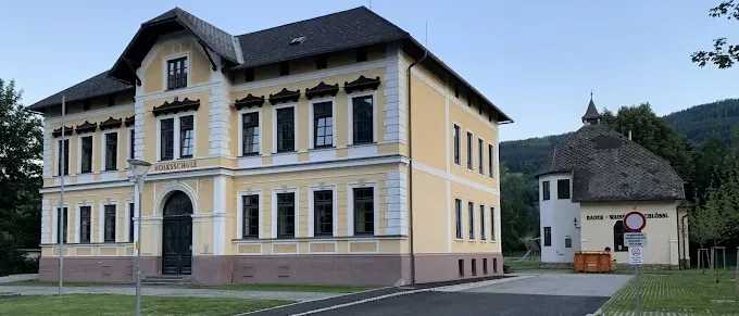 Ein großes gelbes Gebäude mit schwarzem Dach und vielen Fenstern steht in einer Landschaft mit grünem Rasen und Bergen im Hintergrund.